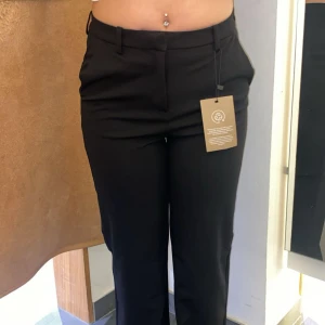 Svarta högmidjade kostymbyxor - Snygga svarta högmidjade kostymbyxor med klassisk design.Byxorna har hällor för bälte och stängs med dragkedja och hake. Perfekta till en stilren look och passar till många olika tillfällen. Med fickor på sidorna 