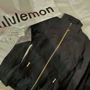 Svart jacka från Lululemon med guldiga dragkedjor - Snygg svart jacka från Lululemon med guldiga dragkedjor framtill och vid fickorna. Figurnära modell med hög krage och långa ärmar. Perfekt för dig som gillar sportig och stilren look.