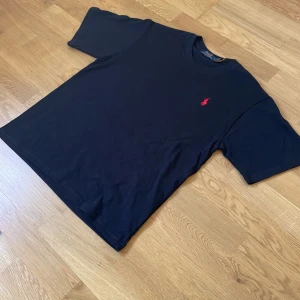 Svart t-shirt från Polo Ralph Lauren - Klassisk svart t-shirt från Polo Ralph Lauren med röd broderad logga på bröstet. T-shirten har rund halsringning och är tillverkad i mjuk bomull. Perfekt basplagg med stilren design och normal passform. Säljer pga var för stor och rev av prislappen för tidigt, hojta till om ni har frågor eller funderingar😃