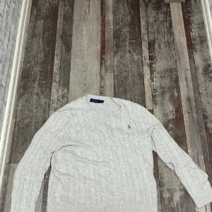  Ralph Lauren stickad tröja  - Säljer en klassisk ljusgrå stickad tröja från Polo Ralph Lauren. Tröjan har kabelstickat mönster, rund halsringning och långa ärmar. 