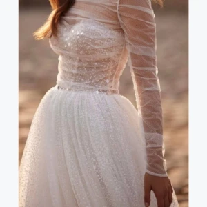 Vit långklänning med glitter och meshdetaljer - Romantisk vit långklänning med glittrigt tyg och transparenta meshärmar. Klänningen har lång ärm och en figurnära överdel med diskreta prickar och vacker struktur. Perfekt för speciella tillfällen.