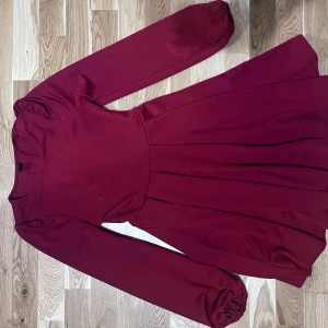 Vinröd långärmad klänning med puffärm - Säljer en vinröd klänning med långa puffärmar och rund halsringning. Klänningen har en utställd kjol och resår vid ärmsluten för en snygg detalj. Perfekt för dig som gillar en enkel men stilren look.  Använd endast på juldagen, mycket fint skick. Totallängd 82 cm  Kommer från djurfritt hem!