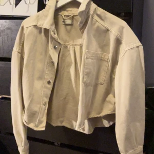 Beige croppad jeansjacka - Säljer en beige croppad jeansjacka med bröstficka och knappar framtill. Jackan har klassisk krage och långa ärmar, perfekt för en trendig och avslappnad look.