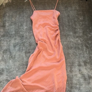 Rosa långklänning med smala axelband från Zara - Säljer en snygg rosa långklänning från Zara med smala axelband och draperad detalj på sidan. Klänningen har en slät, glansig yta och är perfekt för sommarens tillfällen. Enkel och stilren design som känns både trendig och feminin.