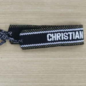 Christian Dior J'ADIOR - Säljer ett svart och vitt armband från Dior J'ADIOR med broderad text och dekorativa tofsar i ändarna. Armbandet har ett flätat band och detaljer i vitt och gult längs kanterna. Perfekt accessoar för att lyfta din stil.