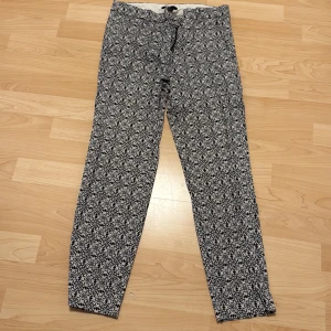 Mönstrade byxor från H&M - Säljer ett par svarta och vita byxor från H&M med ett detaljerat blommigt mönster. Byxorna har normal passform och är tillverkade i ett mjukt bomullsmaterial. Perfekta för dig som vill ha något unikt i garderoben!