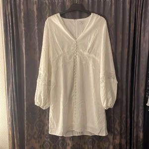 Vit Alicia klänning från Bubbleroom - Supersöt vit klänning från Bubbleroom med v-ringning, dekorativa knappar framtill och spetsdetaljer över byst och ärmar. Långärmad modell med ballongärmar och kort längd. Perfekt för somriga tillfällen eller student.