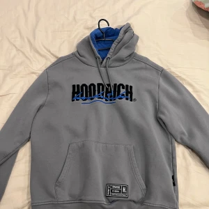 Grå hoodie från Hoodrich med blå detaljer - Säljer en grå hoodie från Hoodrich med svart och blå broderad logga på bröstet och ryggen. Tröjan har huva, magficka och ribbade muddar. Perfekt för en avslappnad stil.