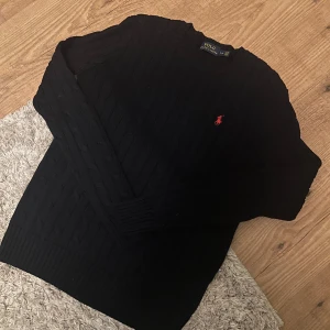 Svart kabelstickad tröja från Polo Ralph Lauren - Kabelstickad svart tröja från Polo Ralph Lauren med rund halsringning och klassisk röd logga broderad på bröstet. Tröjan har lång ärm och ribbade muddar vid ärmslut och nederkant. Perfekt för en stilren och tidlös look.