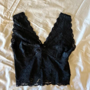 Svart spetsig croppad linnetopp - Säljer en svart croppad linnetopp i spets med djup v-ringning och breda axelband. Toppen har ett vackert blommigt spetsmönster och är perfekt för att lyfta vilken outfit som helst.