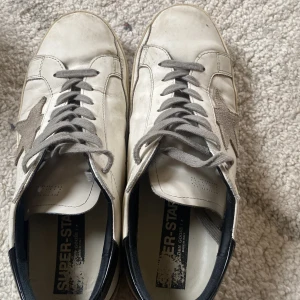 Vita sneakers från Golden Goose med grå stjärna - Säljer ett par vita sneakers från Golden Goose med grå snörning och en grå stjärndetalj på sidan. Skorna har svart hälparti och klassisk rund tå. Kommer med originalkartong.