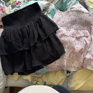 Två volangkjolar från Gina Tricot - Säljer två söta kjolar från Gina Tricot i storlek 158/164. En är helsvart och en är vit med rosa blommigt mönster. Båda har bred smock i midjan och dubbla volanger för extra volym. Perfekta för dig som gillar en feminin och trendig stil.