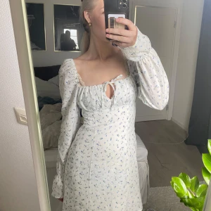 Vit blommig klänning med puffärmar - Supersöt vit klänning med små blå blommor och långa puffärmar. Klänningen har en söt knytning framtill vid bysten och lätt rynkade detaljer vid axlarna. 