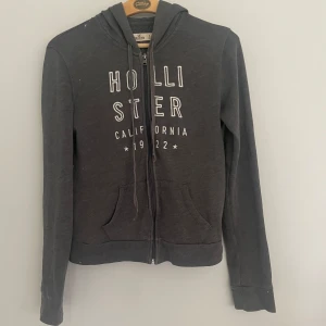 Mörkgrå hoodie från Hollister - Mörkgrå hoodie från Hollister med vit broderad text på bröstet. Tröjan har dragkedja, rymlig luva  och två fickor framtill. 