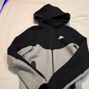 Nike hoodie med dragkedja - Säljer en snygg hoodie från Nike i svart och grått med dragkedja och huva. Jackan har lång ärm och klassisk Nike-logga på bröstet. Perfekt för en sportig och avslappnad stil.
