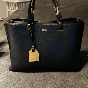 Svart handväska från DKNY - Snygg svart handväska från DKNY i skinn med gulddetaljer och klassisk form. Väskan har två handtag, flera fack inuti och en dekorativ hängande tag. Perfekt för dig som vill ha en stilren accessoar med plats för det viktigaste. Helt oanvänd och skick 10 av 10