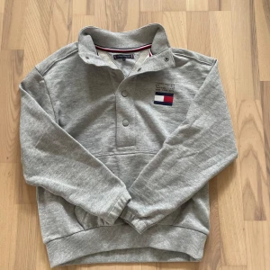 Grå sweatshirt från Tommy Hilfiger - Säljer en grå sweatshirt från Tommy Hilfiger med klassisk logga broderad på bröstet. Tröjan har lång ärm, ribbade muddar och knappar vid halsen. Perfekt för en avslappnad stil.