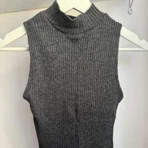 Grå ribbad ärmlös polotopp - Säljer en grå ribbad polotopp. Toppen är ärmlös och har en hög krage som ger en stilren look. Perfekt att styla med jeans eller kjol för en trendig outfit. Använd ett par gånger och är fin och hel! 