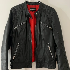 Äkta Svart skinnjacka från Rockandblue - Ursprungspris 2300kr. Äkta svart skinnjacka från Rockandblue med silvriga dragkedjor och röd insida. Jackan har fyra fickor med dragkedja framtill, quiltade axelpartier , baksida och bälte i nederkant. Perfekt för en cool och stilren look.
