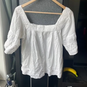 Vit blus med spetsdetaljer från Oysho - Säljer en luftig vit blus från Oysho med vackra spetsdetaljer vid ärmslut, halsringning och nederkant. Blusen har en fyrkantig ringning och trekvartsärmar. Perfekt för varma dagar och ger en romantisk känsla till outfiten.