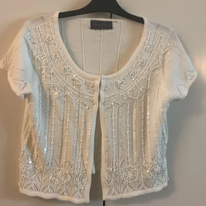 Vit kortärmad mjuk tshirt med paljetter - Superfin vit blustshort med korta ärmar och dekorativa paljetter framtill. Rund halsringning och vackra spetsdetaljer längs nederkanten. Perfekt att slänga över en topp för en extra touch.