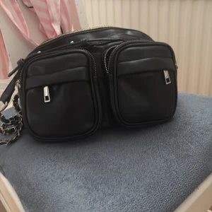 Svart noella väska  - Säljer en svart axelväska med två stora framfickor och silverfärgade dragkedjor. Väskan har en chunky kedja som axelrem och är tillverkad i ett slätt material. Perfekt för att bära det viktigaste med stil.
