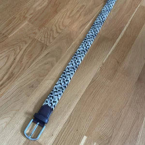 Flätat bälte från ONEWear Sweden - Snyggt flätat bälte i svart, grått och vitt från ONEWear Sweden. Bältet har detaljer i mörkbrunt skinn och ett silvrigt spänne. Perfekt accessoar för att lyfta din outfit.
