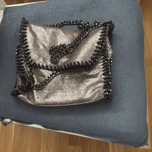 Glittrig silver axelväska med kedjedetaljer - Säljer en snygg glittrig silver axelväska med svarta kedjedetaljer runt kanterna och som axelrem. Perfekt för att lyfta vilken outfit som helst! Väskan har ett lock och är lagom stor för det viktigaste.