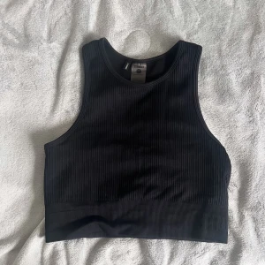 Svart ribbad tränings topp  - Säljer en svart ribbad tränings topp från Stronger. Toppen har en hög halsringning och är ärmlös, perfekt för träning. Enkel och stilren design som passar till mycket. 