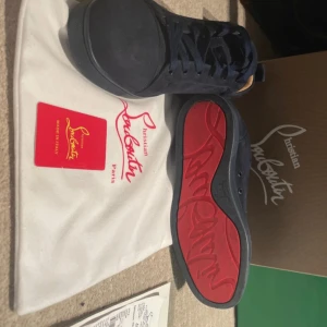 Mörkblå sneakers från Christian Louboutin - Säljer ett par mörkblå sneakers från Christian Louboutin med klassisk röd sula och broderad logga på hälen. Skorna har rund tå, platt sula och är tillverkade i mocka. Snygg och stilren design med svarta skosnören.