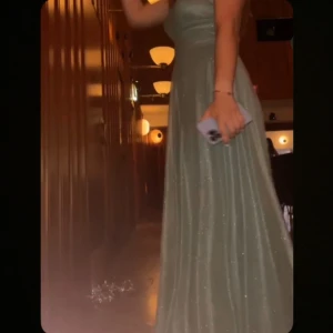 Ljusgrön glittrig balklänning med v-ringning - Säljer en elegant ljusgrön långklänning med glittrigt tyg och v-ringad hals. Klänningen har breda axelband och markerad midja som ger en fin siluett. Perfekt för bal eller festliga tillfällen. 