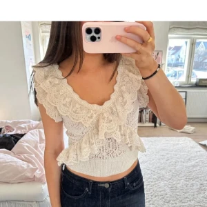 Vit spetsblus med volang - Supersöt vit blus i spets med djup v-ringning och volangdetaljer längs axlar och byst. Toppen är croppad och har ett romantiskt, feminint uttryck. Perfekt att styla med jeans eller kjol.