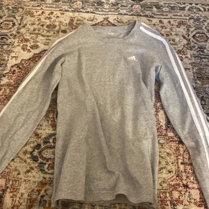 Grå långärmad tröja från Adidas - Säljer en grå långärmad tröja från Adidas med klassiska vita ränder längs ärmarna och liten logga på bröstet. Perfekt för en sportig och avslappnad stil.