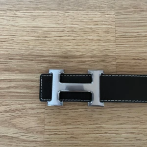 Svart skärp med silverfärgat H-spänne - Säljer ett svart skärp med ett stort, silverfärgat H-spänne. Skärpet är gjort i skinn med vit kontrastsöm längs kanterna. Perfekt accessoar för att lyfta din outfit. Ej använd med har inte prislapp.