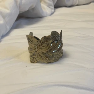 Chucky smycke - Unik och bred armring i guldfärgad metall med detaljerat lövmönster och utskurna hjärtan. Perfekt för dig som vill sticka ut med ett naturinspirerat smycke. 