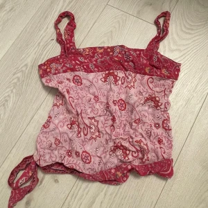 Rosa och röd mönstrad linnetopp - Säljer en söt linnetopp i rosa och rött med blommigt och paisley-inspirerat mönster. Toppen har breda axelband och knyt i sidan samt dekorativa detaljer längs nederkanten. Perfekt för varma dagar! OBS!! Andra bilden är egentligen framsidan. Står inhen storlek men skulle säga ca S eller XS