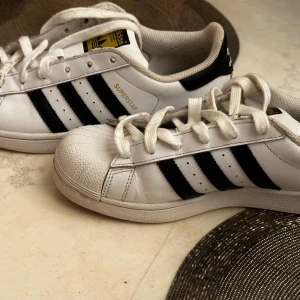 Adidas Superstar vita sneakers med svarta ränder - Klassiska Adidas Superstar sneakers i vitt med svarta ränder och gummitå. Skorna har snörning och den ikoniska Adidas-loggan på hälen och plösen. Perfekta för dig som gillar en tidlös och sportig stil.