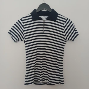 Randig pikétröja från Hennes Collection - Säljer en svartvit randig pikétröja från Hennes Collection. Tröjan har korta ärmar, klassisk krage och dragkedja framtill. Perfekt för en stilren och avslappnad look.