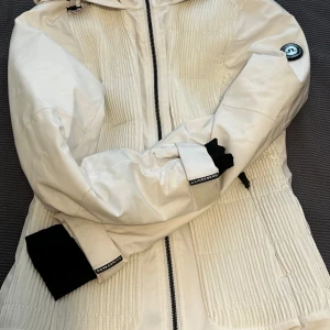 Vit vinterjacka från J.Lindeberg - Snygg vit vinterjacka från J.Lindeberg med svarta detaljer och dragkedja framtill. Jackan har huva, flera fickor med dragkedja och quiltade partier. Perfekt för kalla dagar och har en sportig look med logga på ärmen.