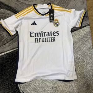 Real Madrid hemmatröja 2023/24 från Adidas - Säljer en officiell Real Madrid hemmatröja från Adidas med gula och mörkblå detaljer. Tröjan har klubbmärket på bröstet, sponsortryck och klassiska Adidas-ränder på axlarna. Tillverkad i lätt och ventilerande material.