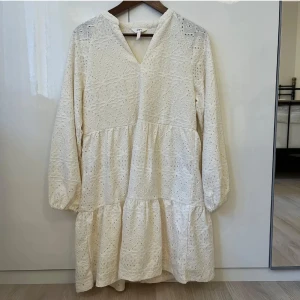 Vit spetsklänning med v-ringning - Söt vit klänning med v-ringning och långa ärmar. Klänningen har broderade spetsdetaljer och en luftig, volangig kjol. Perfekt för sommarens tillfällen och midsommarfirande.