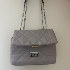 Grå quiltad axelväska med kedjerem - Snygg grå axelväska med quiltat mönster och silverfärgad kedjerem. Väskan har ett lock med metallspänne och ett innerfack med dragkedja. Perfekt för att bära det viktigaste med stil.(pris går diskutera)