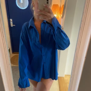 Oversized blå skjorta - Säljer en oversized blå skjorta med klassisk krage och knäppning framtill. Skjortan har långa ärmar och en avslappnad passform, perfekt att styla lager på lager eller bära som den är. Använt som strand skjorta över bikinin, så fint!