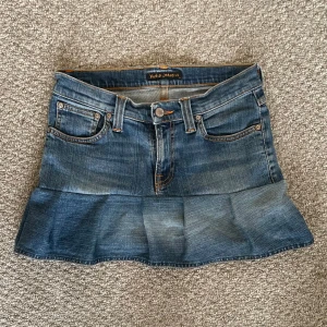 Jeanskjol med volang från Nudie Jeans Co - Säljer en blå jeanskjol från Nudie Jeans Co med volang nertill. Kjolen har klassiska jeansfickor fram och bak, orangea kontrastsömmar, knappstängning framtill och detaljer på bakfickorna. 
