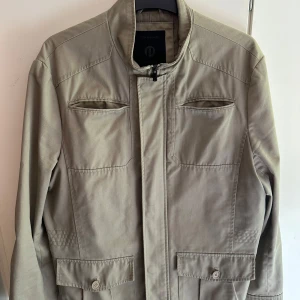 Beige jacka från Jack & Jones - Stilren beige jacka från Jack & Jones med ståkrage och dragkedja framtill. Jackan har två bröstfickor med dragkedja och två fickor med lock och knapp nertill. Perfekt för vår och höst. En knapp saknas vid handleden. 