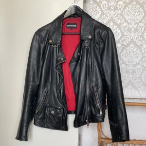 Svart skinnjacka från Rockandblue - Säljer en klassisk svart skinnjacka av lammskinn från Rockandblue med röd insida. Jackan har flera dragkedjor, knappar vid axlarna och detaljer vid midjan. Enda slitaget är på bilden till höger.