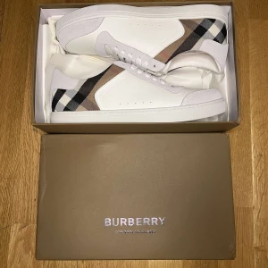 Burberry vita sneakers med rutigt mönster - Säljer ett par snygga vita sneakers från Burberry med klassiskt beige, svart och brunt rutigt mönster på sidorna. Skorna har snörning, rund tå och platt sula. Perfekta för dig som vill ha en stilren och lyxig look.