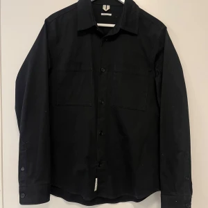 Svart overshirt från arket med bröstfickor - Säljer en svart overshirt från arket med klassisk krage och två bröstfickor. Skjortan har knäppning framtill och långa ärmar. Passformen är regular fit och materialet känns som bomull. Perfekt för en stilren look.