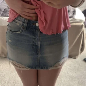 Blå jeanskjol med slitna detaljer - Säljer en kort blå jeanskjol med slitna kanter och fransar längst ner. Kjolen har fickor fram och bak samt knapp och dragkedja framtill.