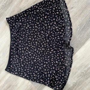 Svart blommig kjol från H&M  - Säljer en svart blommig kjol från H&M med små rosa och gröna blommor. Kjolen har en diskret dragkedja i sidan och har extra tyg under. Perfekta för varma dagar och har en tajt passform och lite utsväng längst ner 